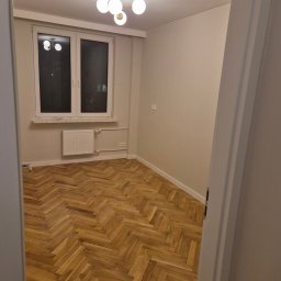 MARKS-BUD - Pusty pokój po remoncie z parkietem ułożonym w jodełkę, widoczne okno, grzejnik i nowoczesna lampa sufitowa z białymi kulami.