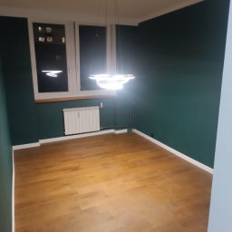 MARKS-BUD - Pusty pokój z drewnianą podłogą, ciemnozielonymi ścianami i nowoczesną, okrągłą lampą wiszącą z trzema pierścieniami LED.