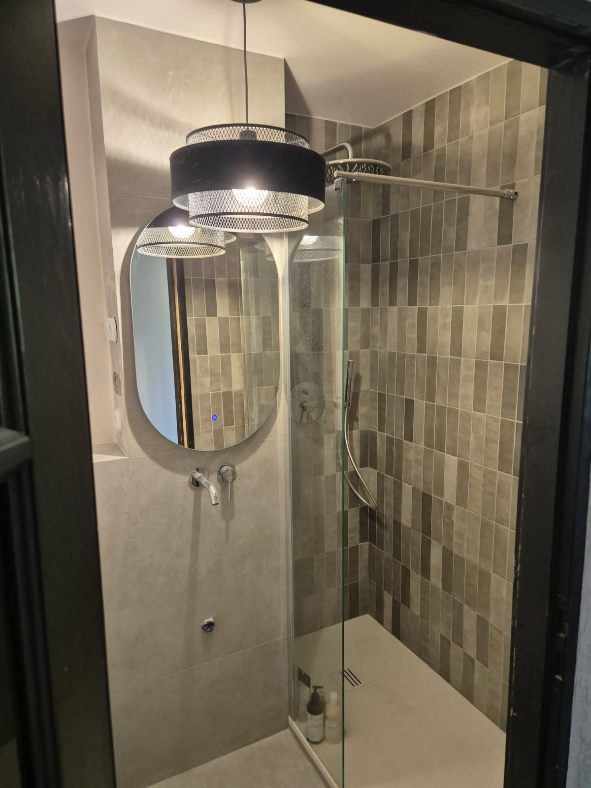 Nowoczesna łazienka z szarą, pionową glazurą, kabiną prysznicową ze szklaną ścianką, owalnym lustrem z podświetleniem LED i designerską lampą wiszącą z metalową siatką.