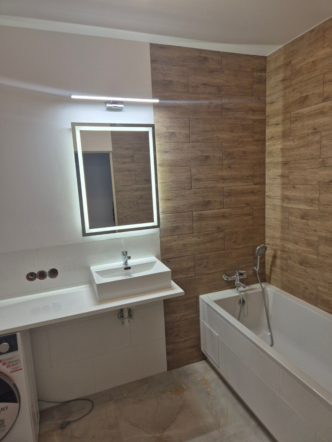 Nowoczesna łazienka po remoncie z białymi płytkami na ścianie, drewnopodobnymi płytkami na części ściany, prostokątną umywalką nablatową, podświetlanym lustrem i białą wanną.