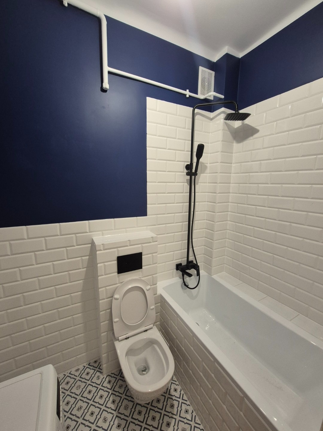 Nowoczesna łazienka z granatową ścianą, białą armaturą i geometryczną podłogą. Widoczna wanna z czarną armaturą natryskową, podwieszana toaleta z otwartą klapą i białe rury na ścianie.