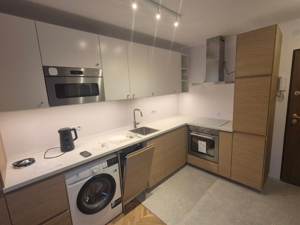 Nowoczesna kuchnia z białymi i drewnianymi szafkami, zintegrowana pralka Electrolux pod blatem, czajnik elektryczny na blacie, oświetlenie podszafkowe LED.