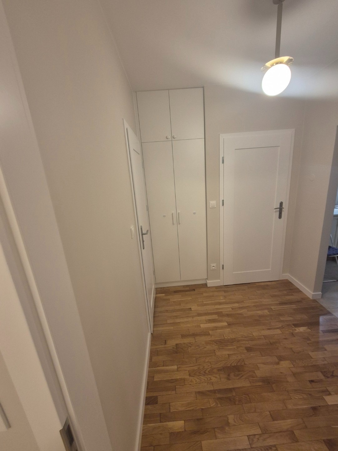 Wnętrze przedpokoju po remoncie z drewnianą podłogą ułożoną w jodełkę, białe drzwi i szafa w zabudowie, oświetlone lampą sufitową.
