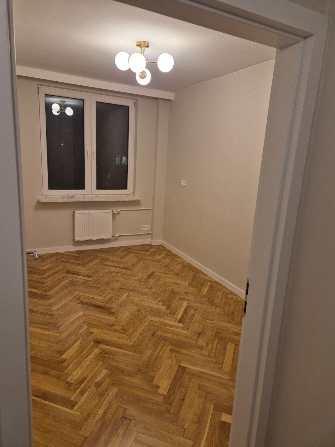 Pusty pokój po remoncie z parkietem ułożonym w jodełkę, widoczne okno, grzejnik i nowoczesna lampa sufitowa z białymi kulami.