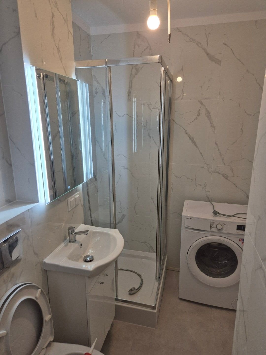 Nowoczesna łazienka z kabiną prysznicową, umywalką z szafką, pralką i marmurowymi płytkami na ścianach, oświetlona żarówką bez klosza.