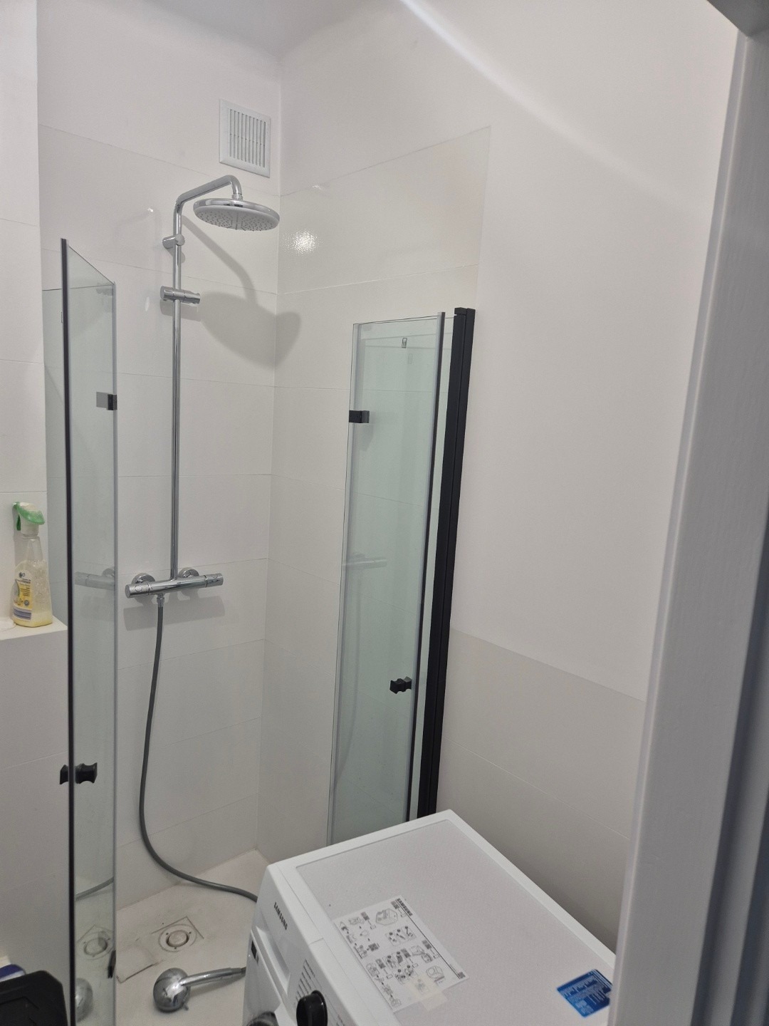 Biała łazienka w trakcie prac wykończeniowych, widoczna kabina prysznicowa typu walk-in z chromowaną deszczownicą, częściowo zamontowana obok pralki Samsung.