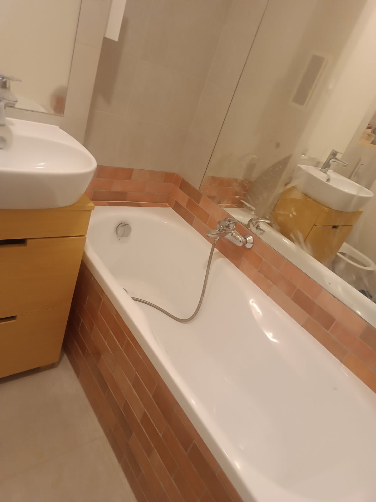 Biała wanna obudowana ceglastymi płytkami, umywalka z szafką w kolorze jasnego drewna, bateria wannowa z wężem, widok w lustrze na toaletę.