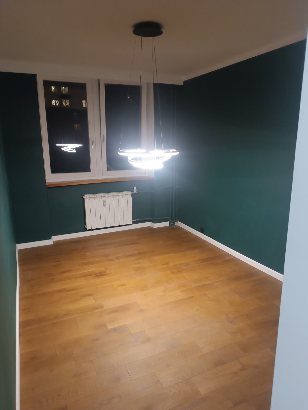 Pusty pokój z drewnianą podłogą, ciemnozielonymi ścianami i nowoczesną, okrągłą lampą wiszącą z trzema pierścieniami LED.