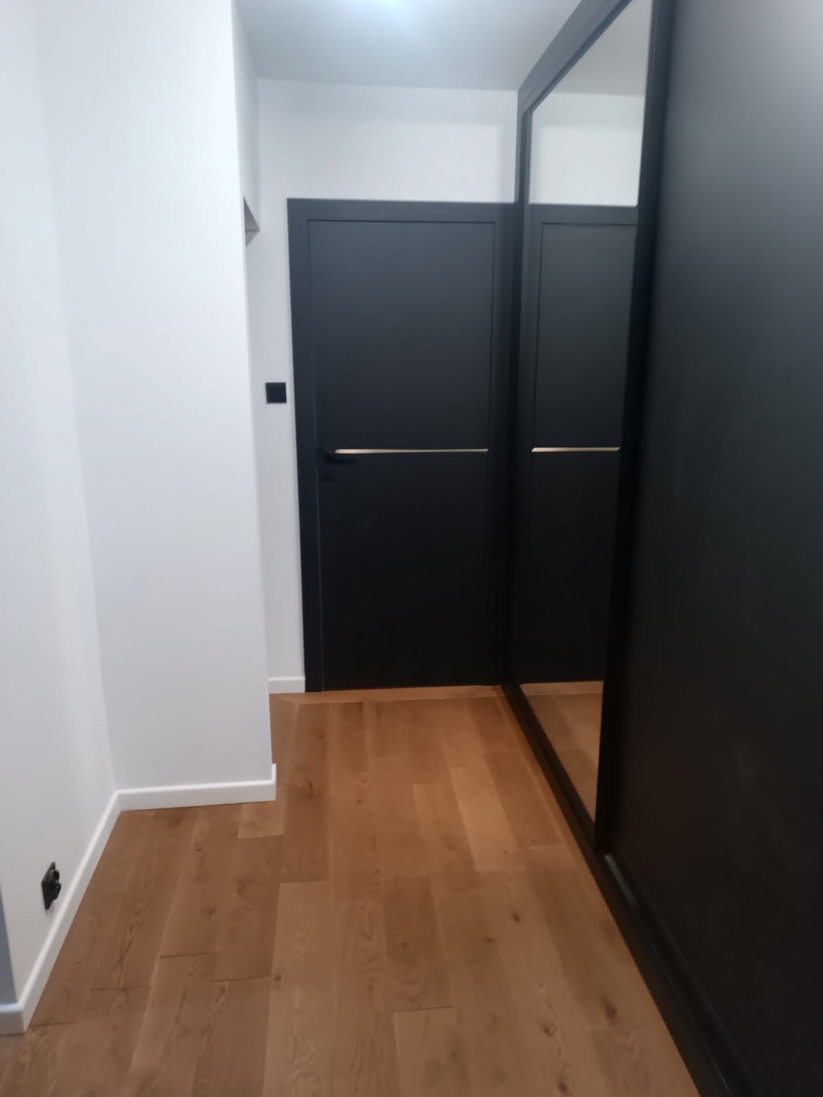 Nowoczesny przedpokój z drewnianą podłogą, białe ściany i czarne drzwi z poziomymi, złotymi uchwytami, oraz szafa z lustrzanymi drzwiami.