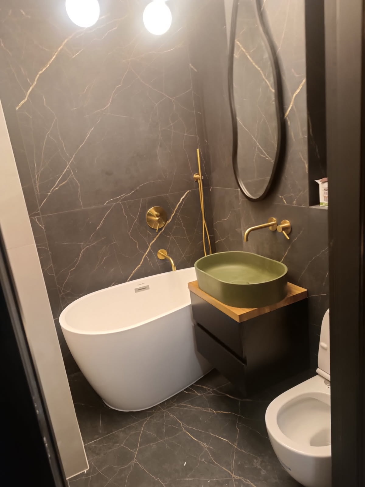 Nowoczesna łazienka z wolnostojącą białą wanną, umywalką w kolorze oliwkowym na drewnianym blacie i czarną szafką, armatura w kolorze złotym, ściany i podłoga wyłożone ciemnym marmurem z jasnymi...
