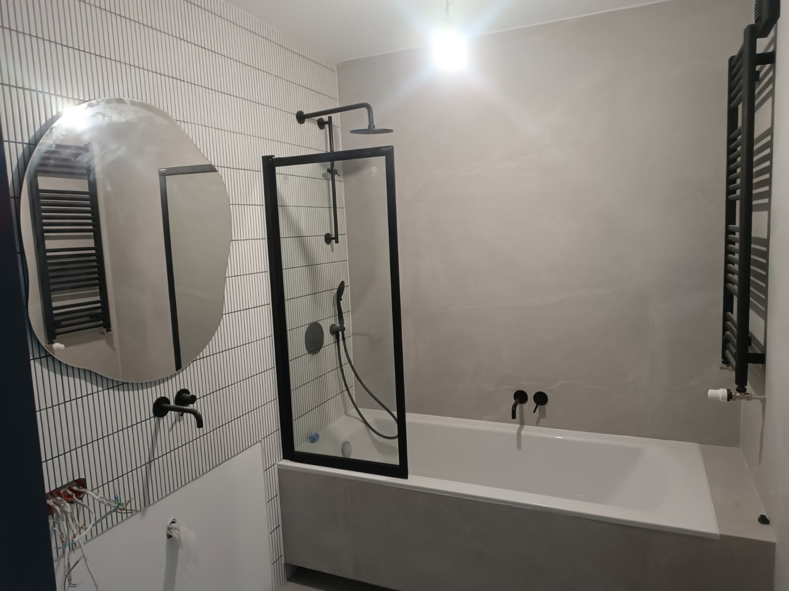 Nowoczesna łazienka z wanną, czarną armaturą i asymetrycznym lustrem, ściany wyłożone białymi, wąskimi płytkami.