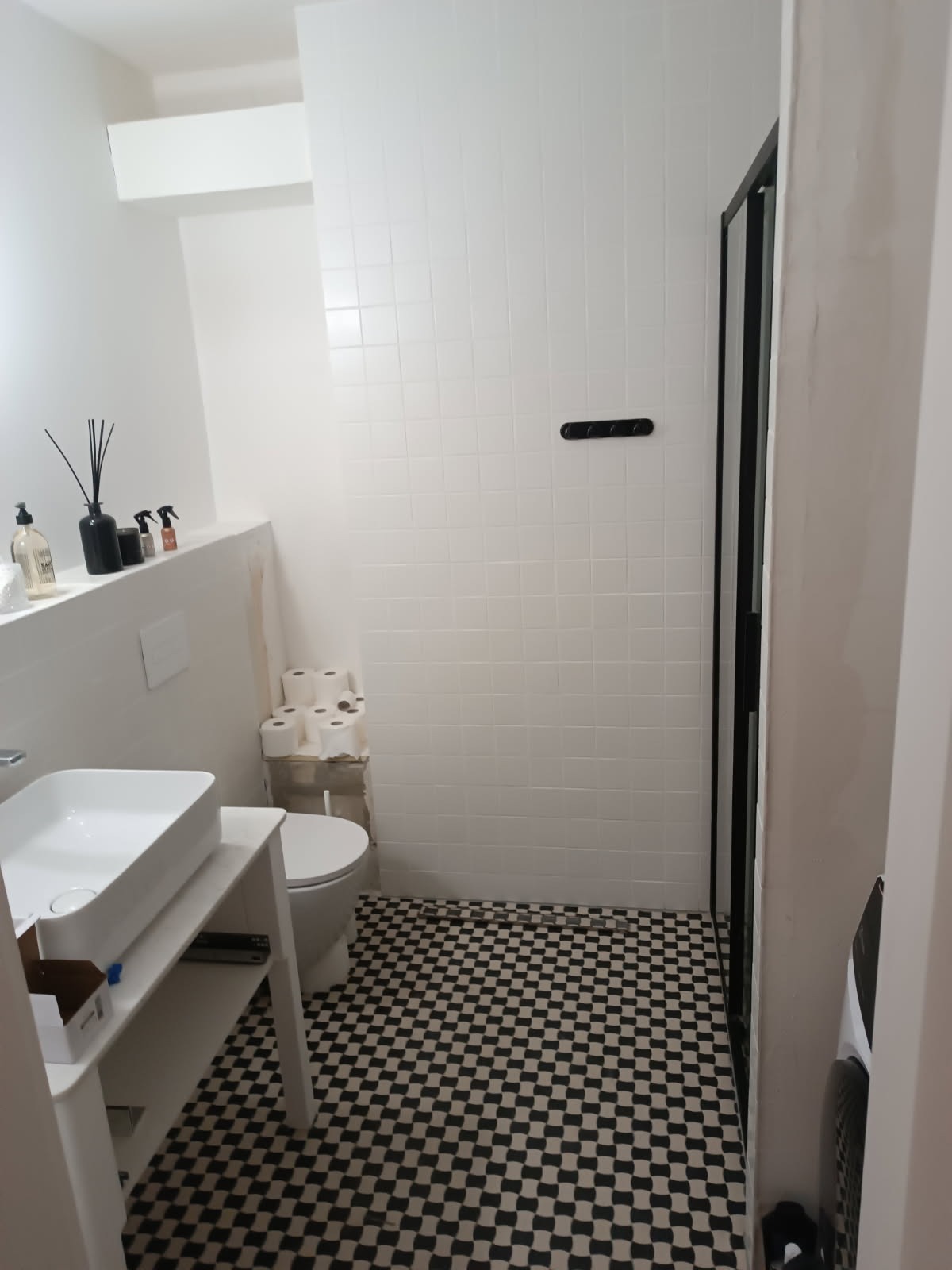 Wnętrze łazienki z białą armaturą, czarno-białą podłogą w szachownicę i białymi płytkami ściennymi. Widoczne niedokończone prace wykończeniowe przy toalecie.