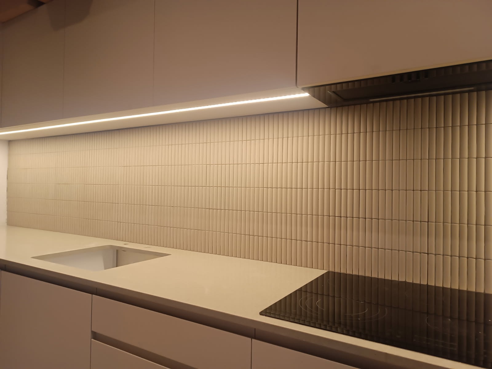 Beżowa kuchnia z płytkami ułożonymi pionowo na ścianie, podświetlenie LED pod szafkami, płyta indukcyjna i wbudowany zlew.