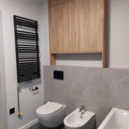 Nowoczesna łazienka z wiszącą toaletą i bidetem, grzejnikiem drabinkowym w kolorze czarnym oraz szafką wiszącą z drewnopodobnym frontem.