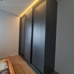 Antima - Czarna szafa wnękowa z przesuwnymi drzwiami i oświetleniem LED w górnej części, widoczny fragment łóżka z beżową narzutą.