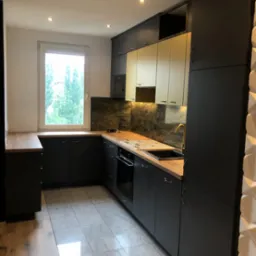 Nowoczesna kuchnia w odcieniach szarości i bieli, z drewnianym blatem i marmurowym backsplashiem, złota bateria, oświetlenie punktowe i dekoracyjna ściana z trójwymiarową fakturą.