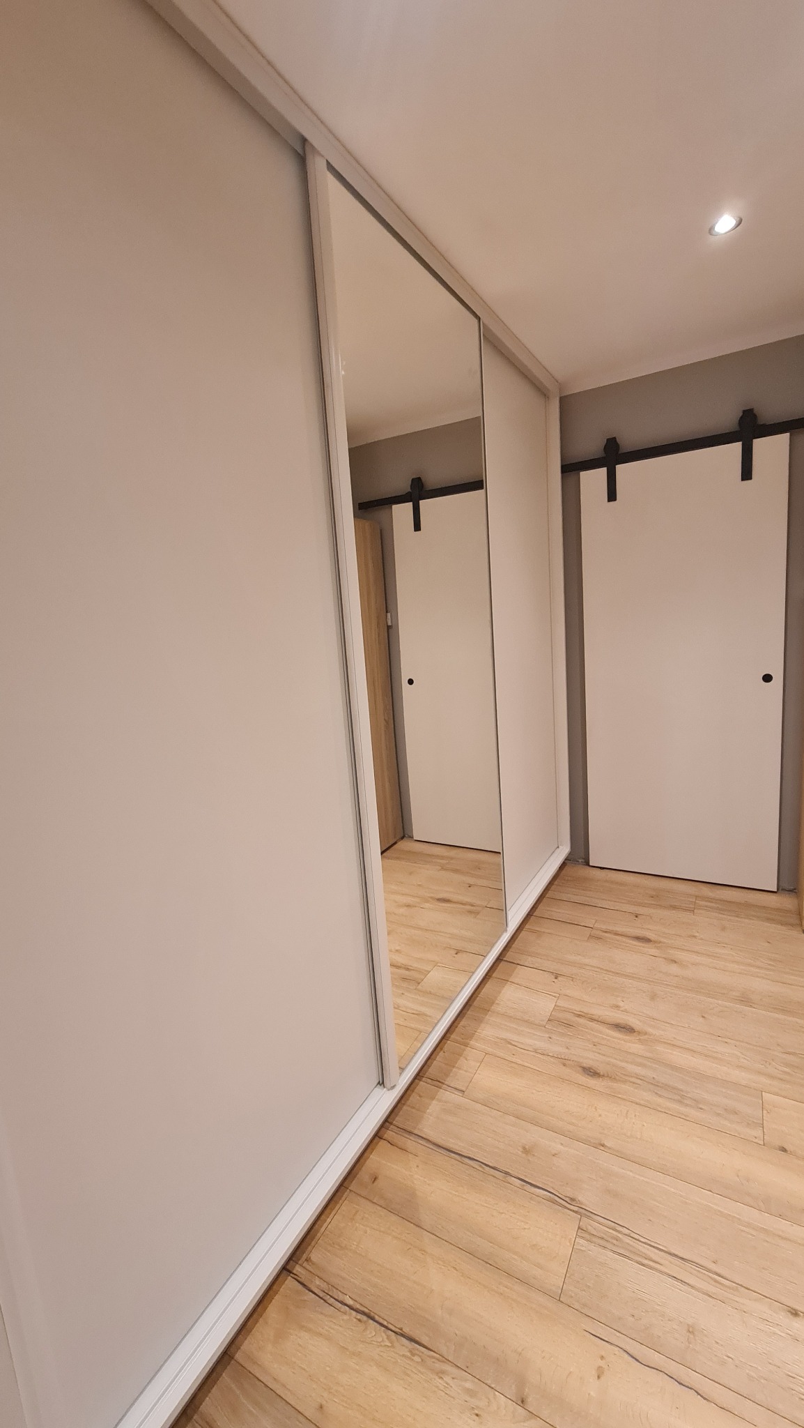 Biała szafa przesuwna z lustrem na całej powierzchni drzwi, widok na podłogę z paneli i drzwi przesuwne w tle.