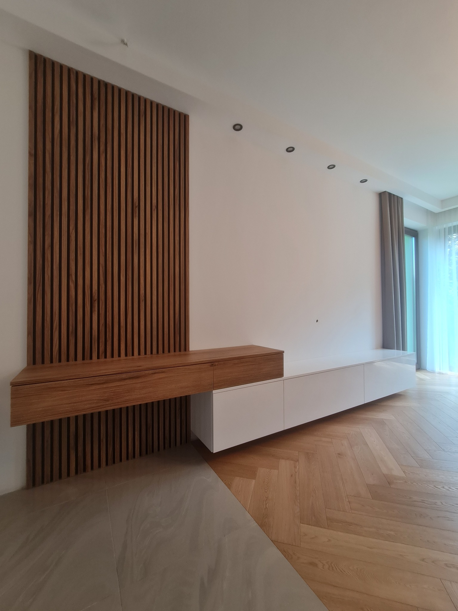 Nowoczesna zabudowa ściany z drewnianymi lamelami i szafką wiszącą z forniru, połączona z białą, niską szafką RTV na parkiecie w jodełkę.
