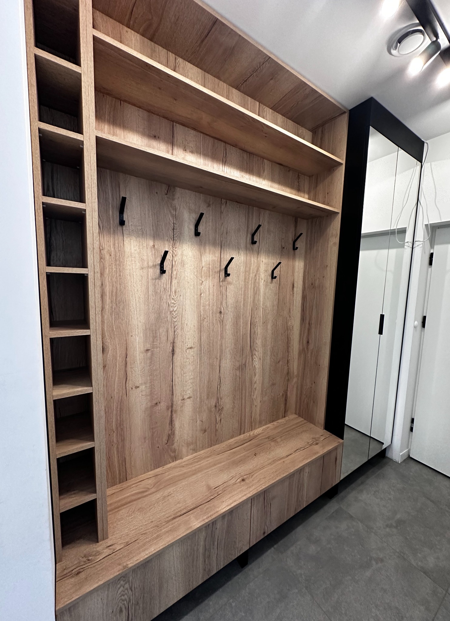 Wnękowa szafa z siedziskiem i otwartymi półkami, wykończona drewnopodobną okleiną w odcieniach beżu i brązu, z czarnymi haczykami na ubrania i lustrem na drzwiach przesuwnych.