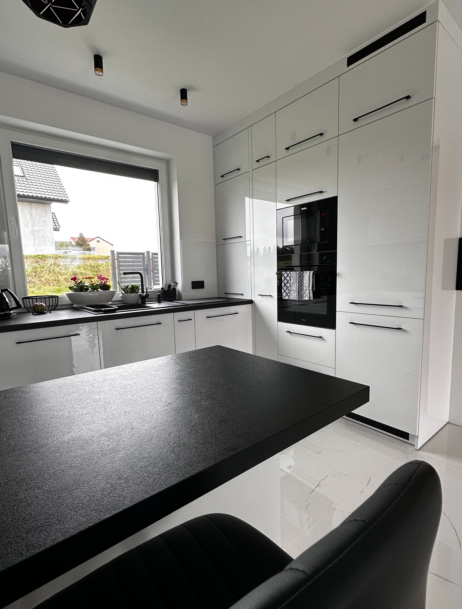 Nowoczesna, biała kuchnia z czarnymi akcentami, w tym czarny blat wyspy i armatura, z widokiem na zewnątrz przez okno.