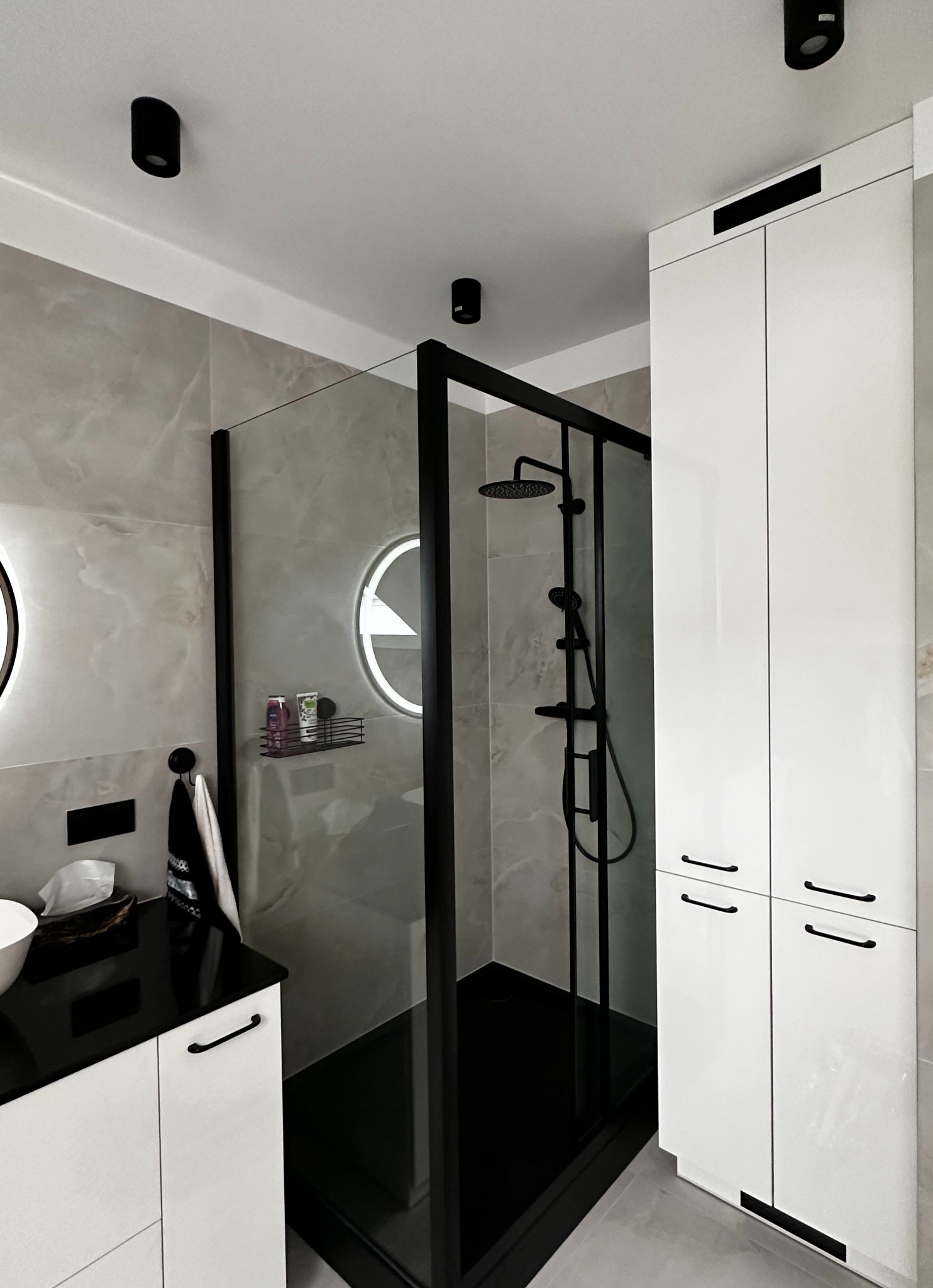 Nowoczesna łazienka z kabiną prysznicową typu walk-in z czarnymi profilami, białe szafki z czarnymi uchwytami, okrągłe lustro z podświetleniem LED i szare płytki imitujące marmur.