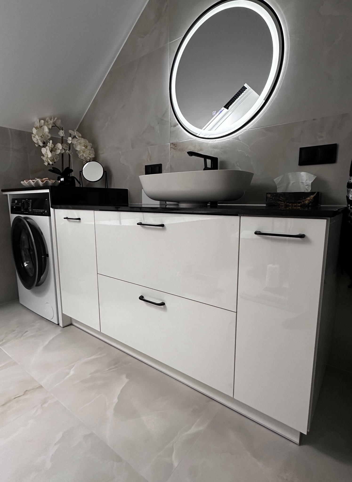 Nowoczesna łazienka na poddaszu z białą szafką pod umywalkę z czarnym blatem, okrągłym lustrem z podświetleniem LED i pralką obok.