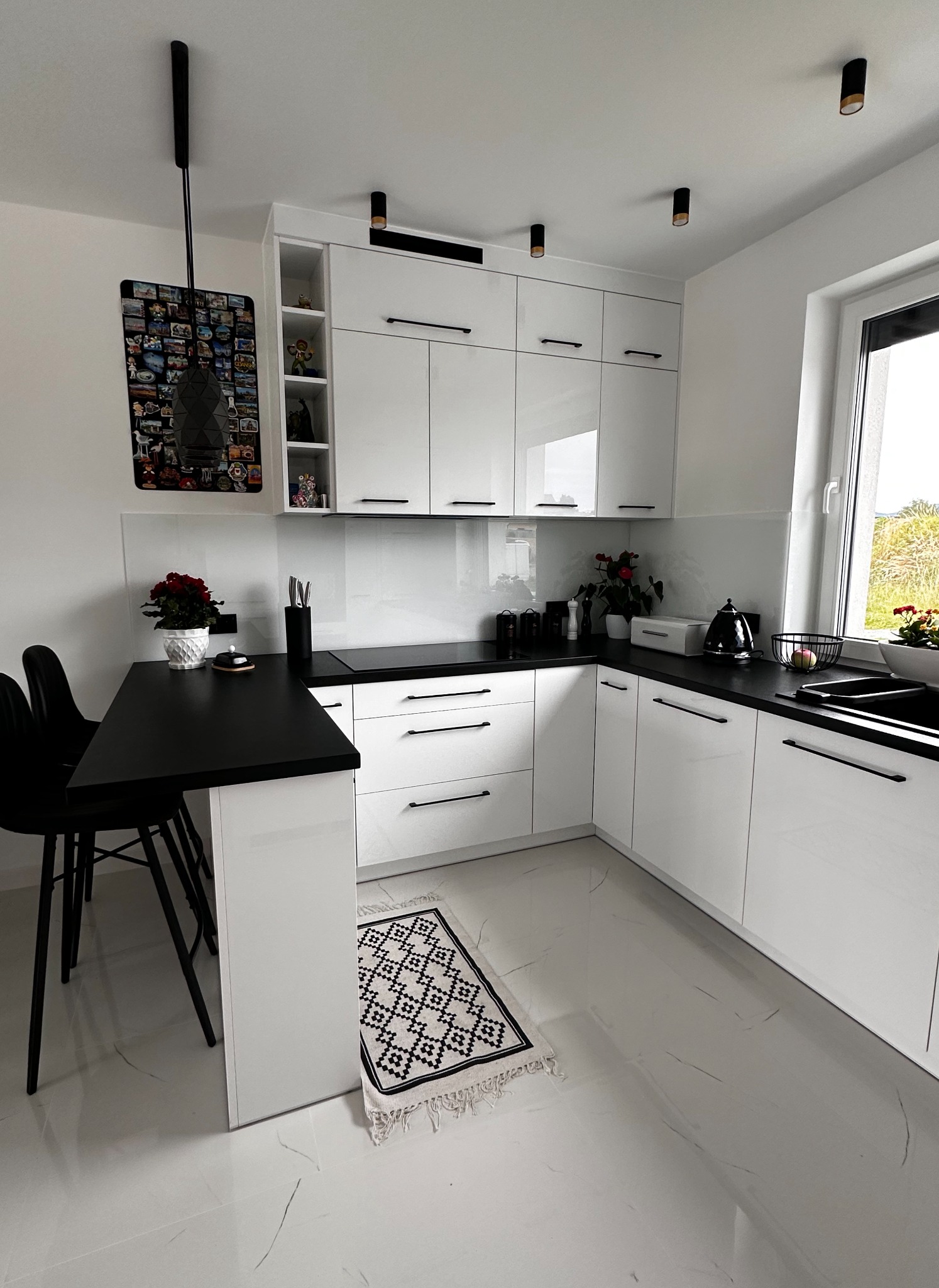 Nowoczesna, biała kuchnia z czarnymi akcentami: blat, uchwyty szafek, krzesła barowe i dywanik z geometrycznym wzorem.