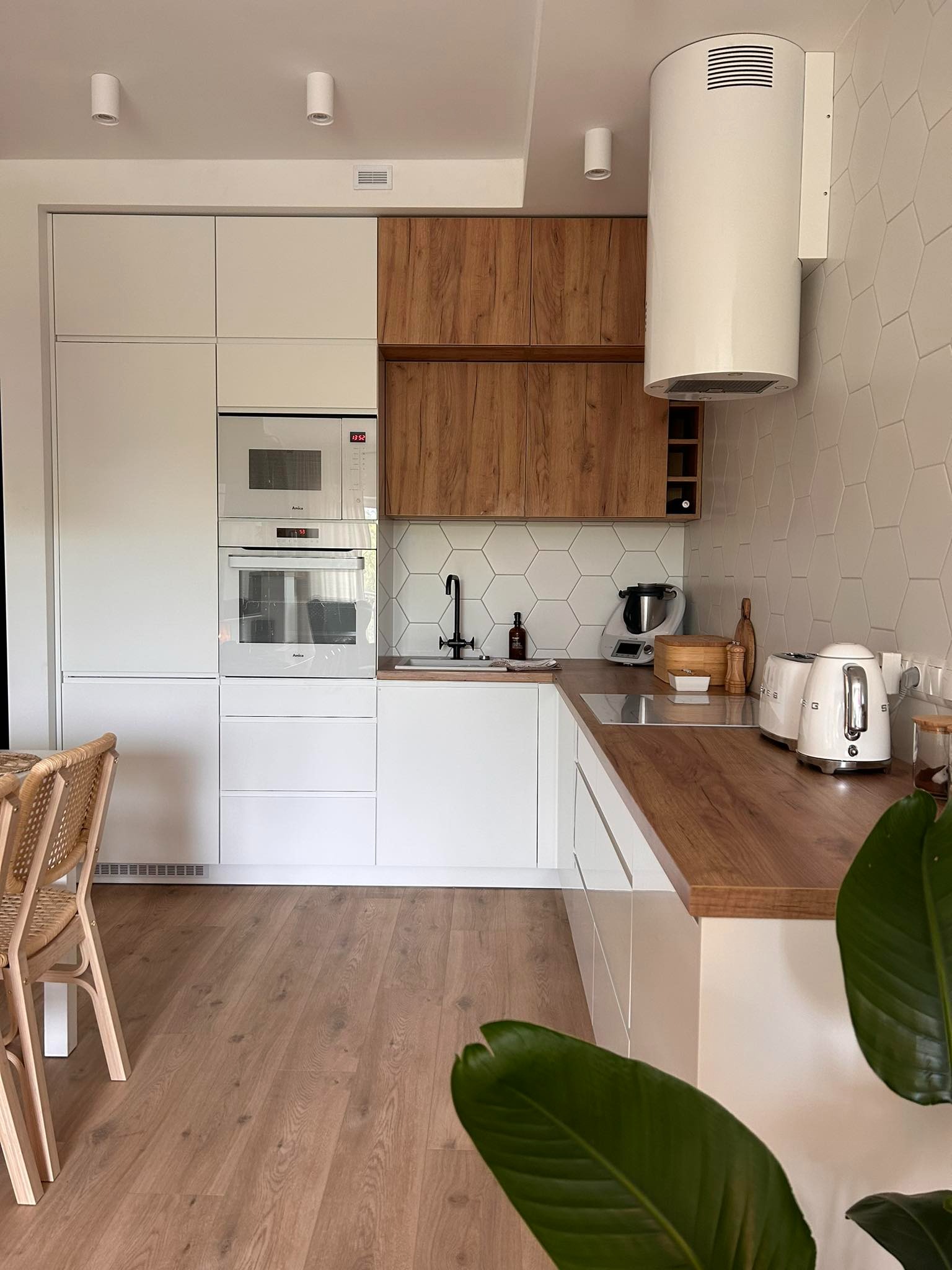 Nowoczesna kuchnia z białymi frontami i drewnianym blatem, uzupełniona o sprzęt AGD w zabudowie oraz heksagonalne płytki na ścianie, z widocznym fragmentem stołu z krzesłami z rattanu.