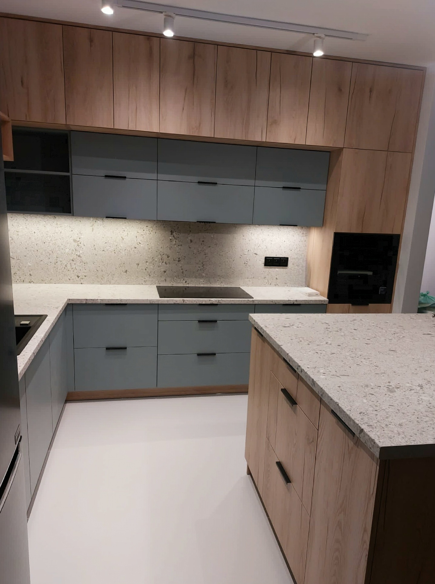 Nowoczesna kuchnia z szarymi szafkami i drewnianymi akcentami, oświetleniem punktowym i blatem z jasnego kamienia.