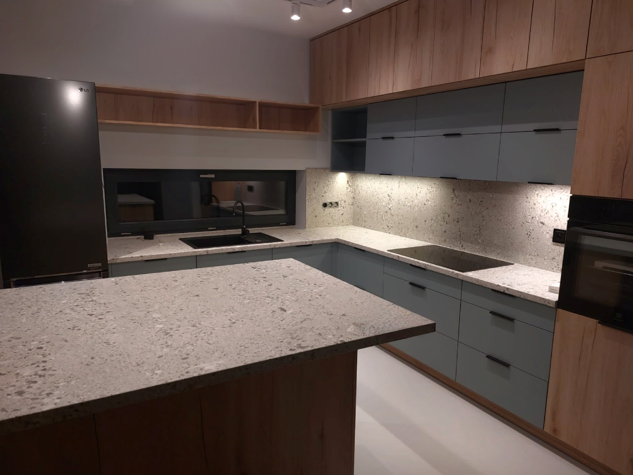 Nowoczesna kuchnia z wyspą, jasnoniebieskie fronty szafek, drewniane wykończenia górnych szafek i wyspy, blaty z szarego kamienia z widoczną fakturą, czarny zlew pod oknem.