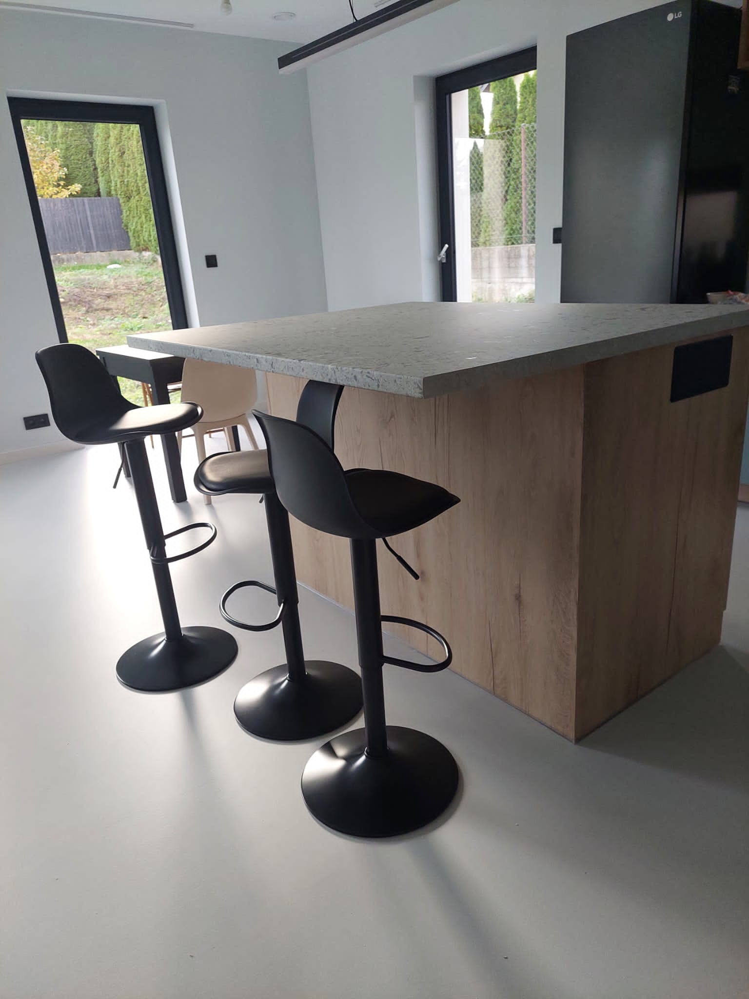 Nowoczesna wyspa kuchenna z blatem w odcieniach szarości i drewnianą obudową, otoczona trzema czarnymi hokerami na jednej nodze. W tle widoczne okno z zielenią i minimalistyczna zabudowa kuchenna.