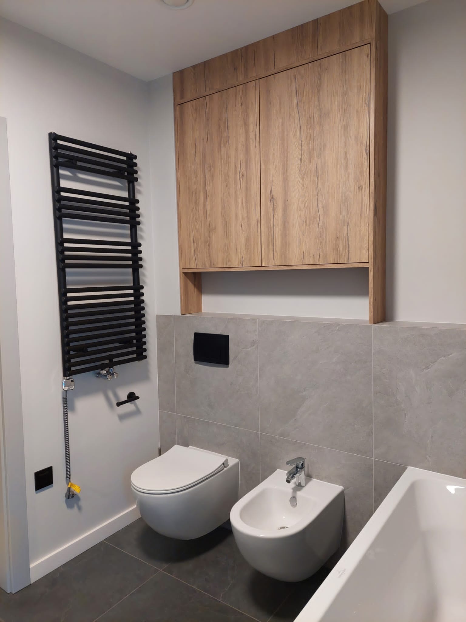 Nowoczesna łazienka z wiszącą toaletą i bidetem, grzejnikiem drabinkowym w kolorze czarnym oraz szafką wiszącą z drewnopodobnym frontem.