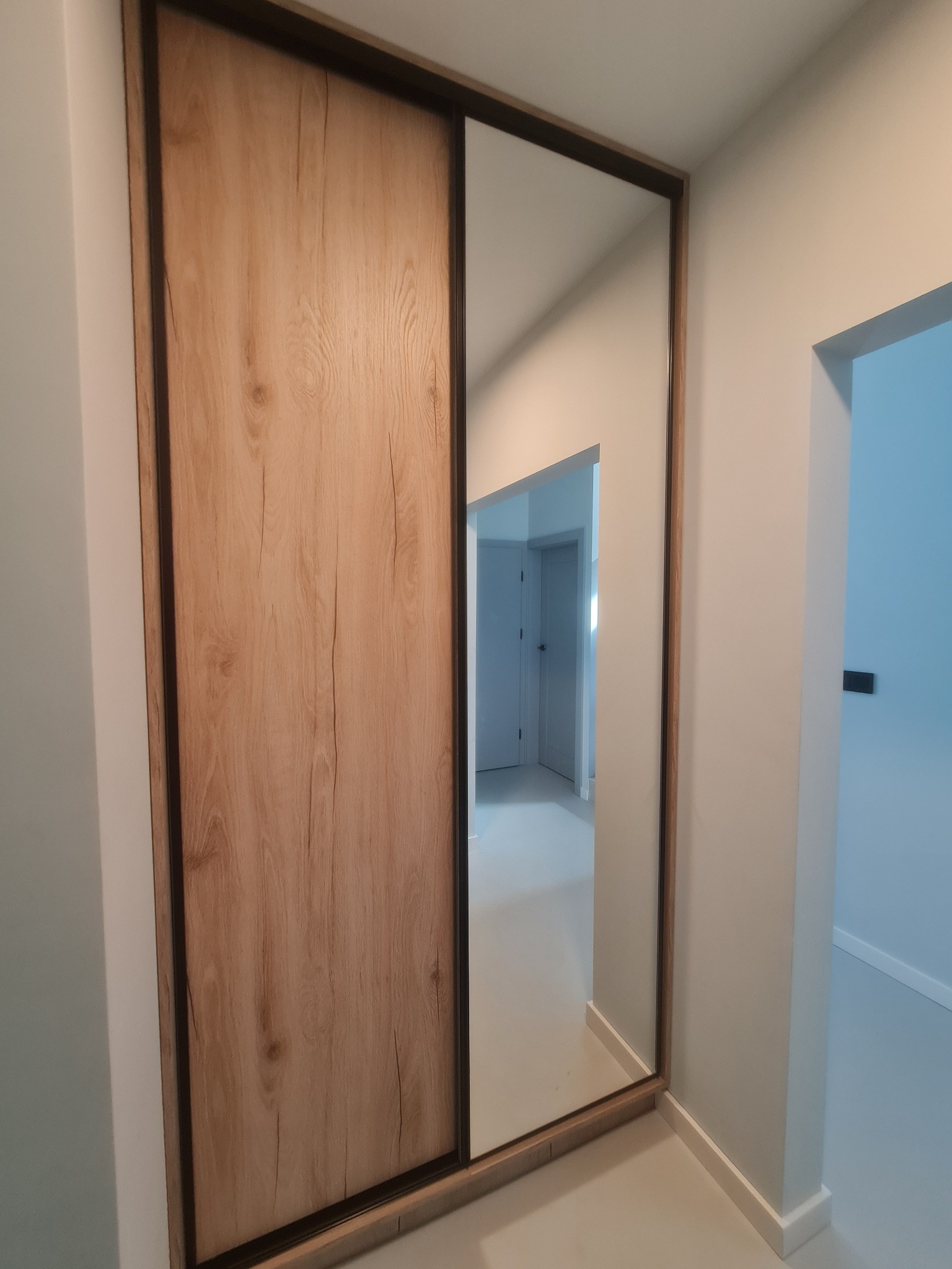 Szafa wnękowa z przesuwnymi drzwiami imitującymi drewno i lustrem, odbijająca fragment wnętrza mieszkania.