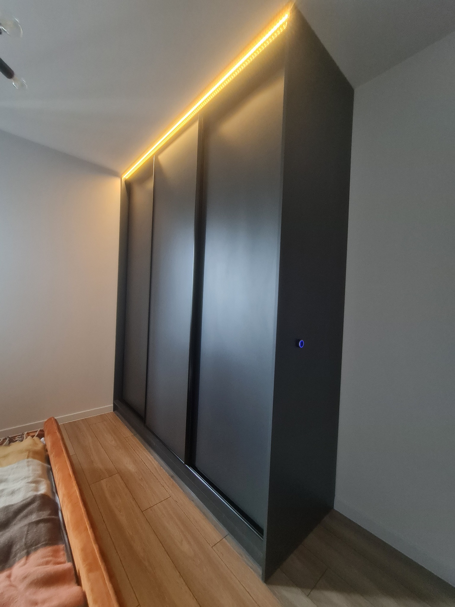 Czarna szafa wnękowa z przesuwnymi drzwiami i oświetleniem LED w górnej części, widoczny fragment łóżka z beżową narzutą.