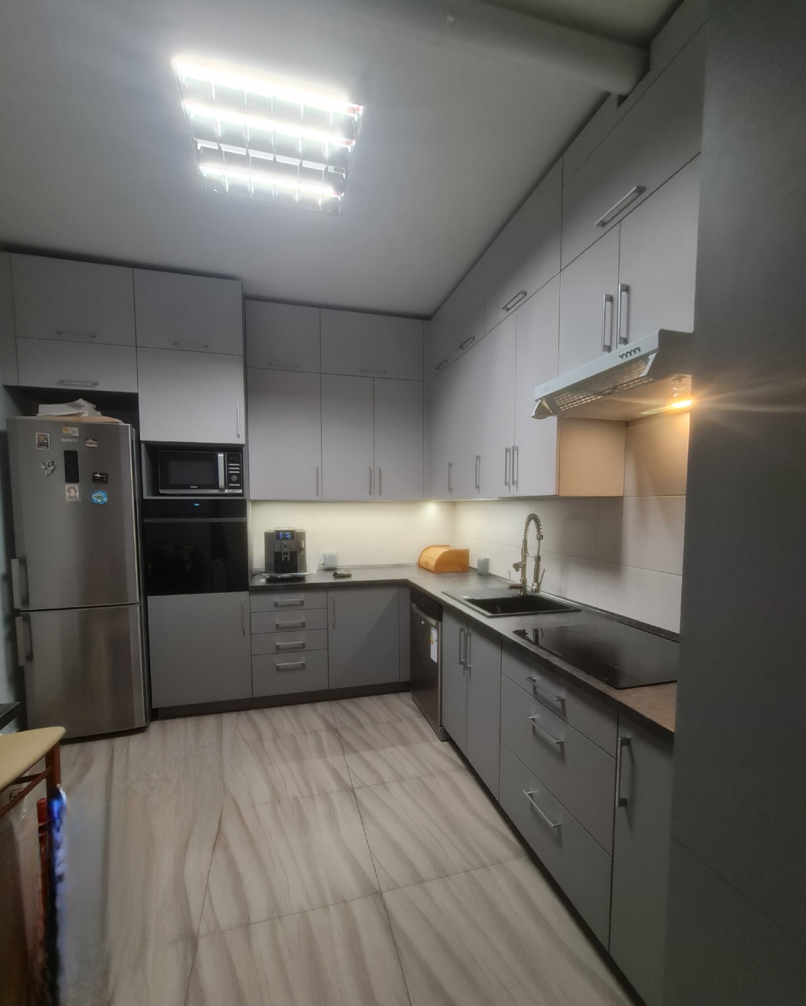 Nowoczesna, szara kuchnia z zabudową pod sufit, oświetleniem LED pod szafkami i płytkami imitującymi drewno na podłodze. Widoczny ekspres do kawy i chlebak na blacie.