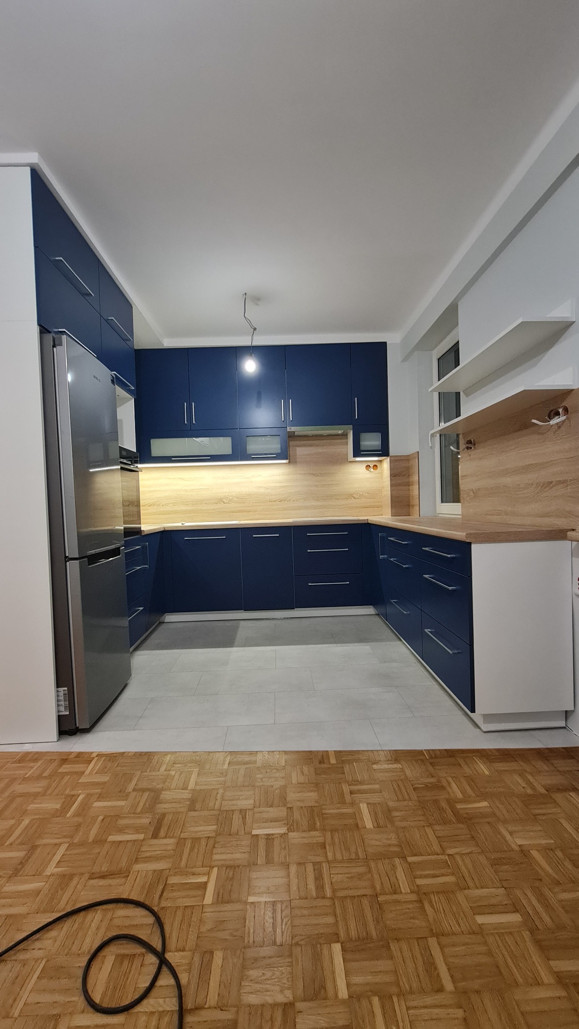 Nowoczesna, granatowa zabudowa kuchenna z drewnianym blatem i srebrną lodówką, widoczna na tle szarej podłogi i parkietu, z oświetleniem podszafkowym i wiszącą żarówką.