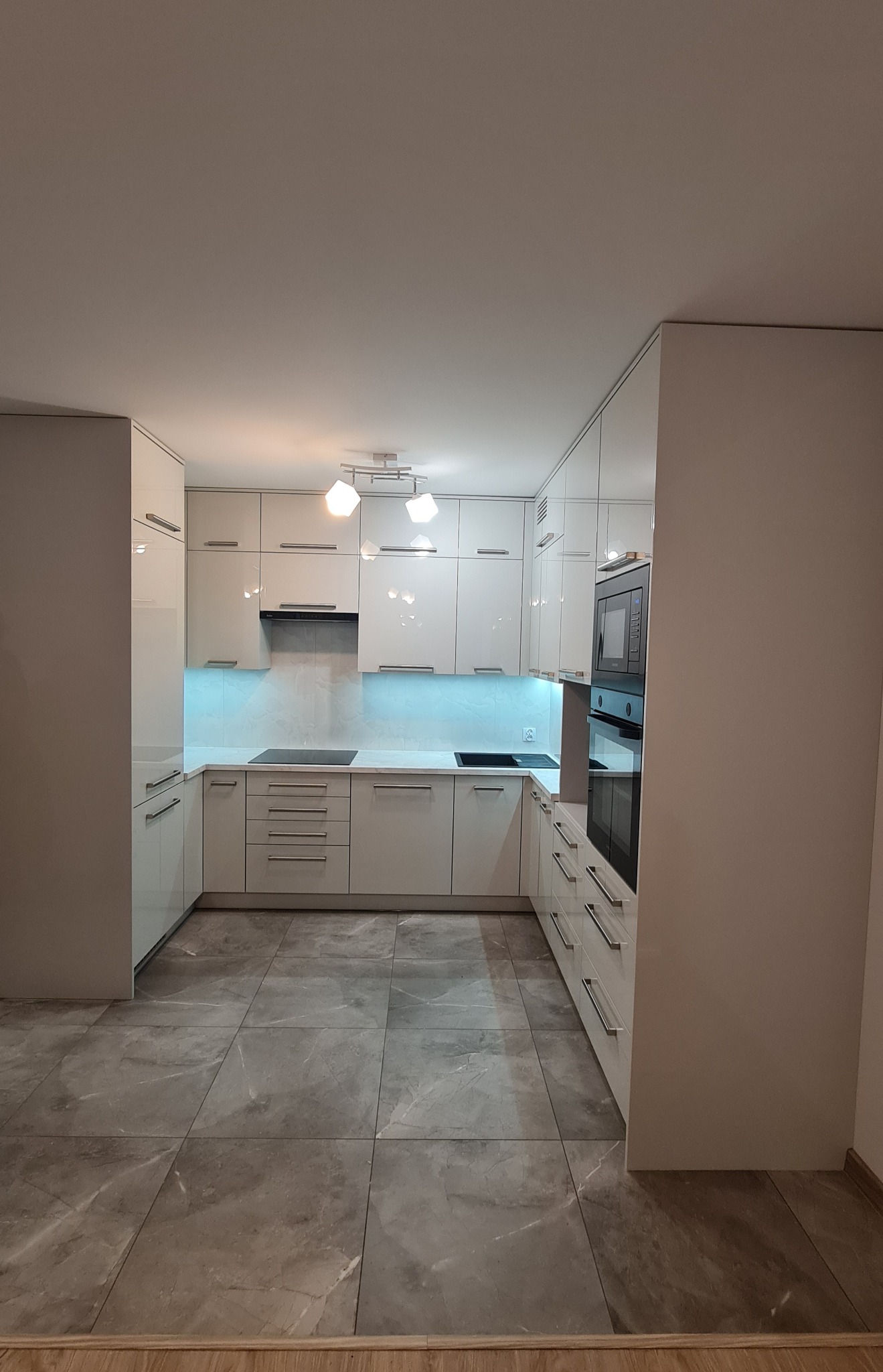 Nowoczesna, biała kuchnia z błyszczącymi frontami, oświetleniem LED pod szafkami i szarą podłogą z płytek imitujących marmur.