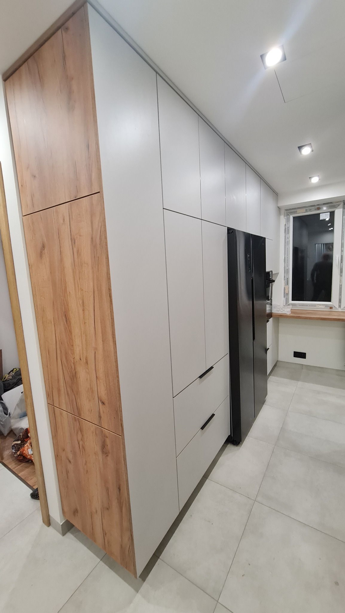 Nowoczesna kuchnia: zabudowa szaf w kolorze białym i drewnopodobnym, zintegrowana z czarną lodówką, minimalistyczne uchwyty, płytki podłogowe w odcieniach szarości.