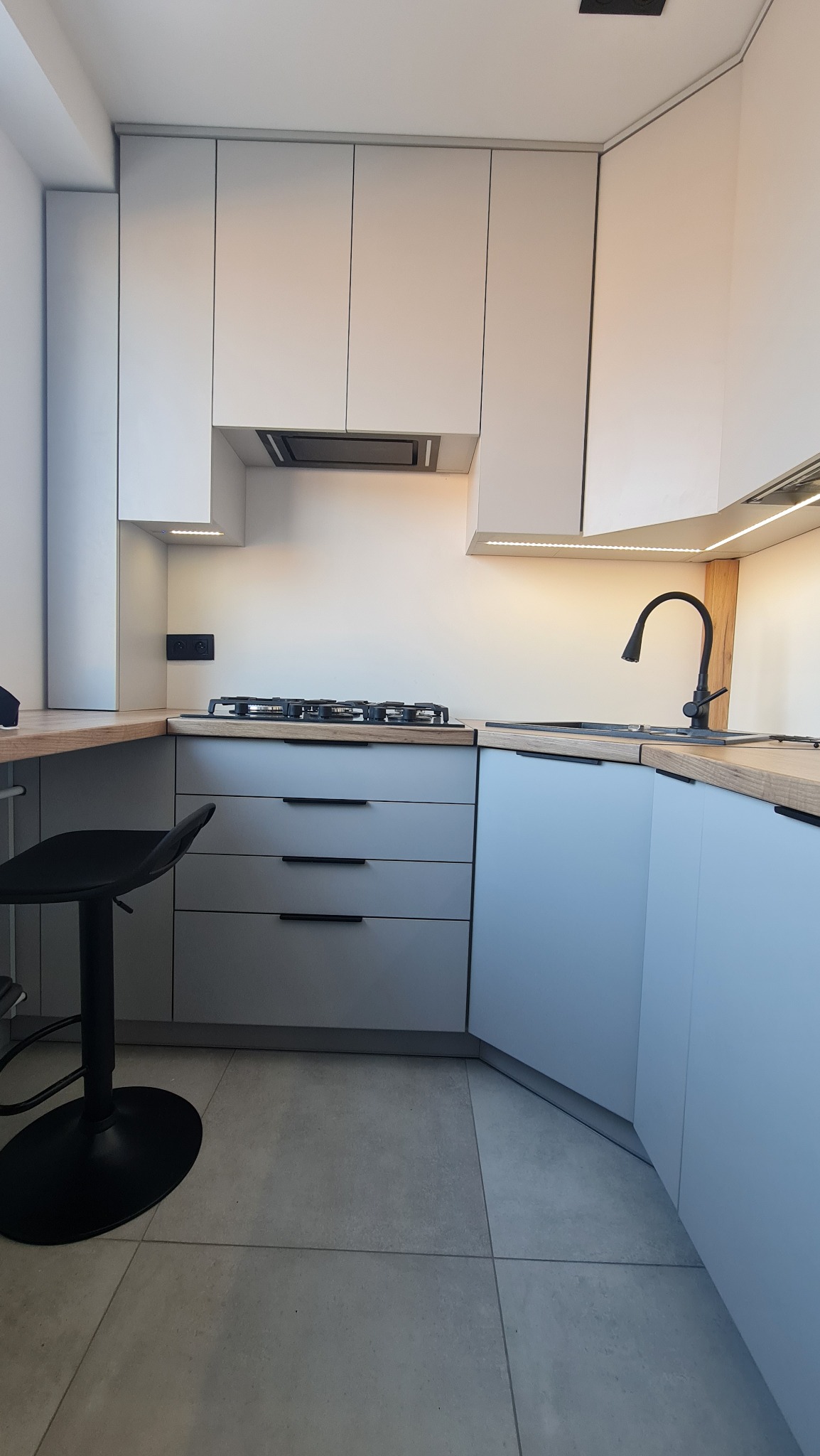Nowoczesna kuchnia z szarymi szafkami, drewnianym blatem i czarną armaturą, oświetlenie LED podszafkowe, widoczny fragment stołka barowego.
