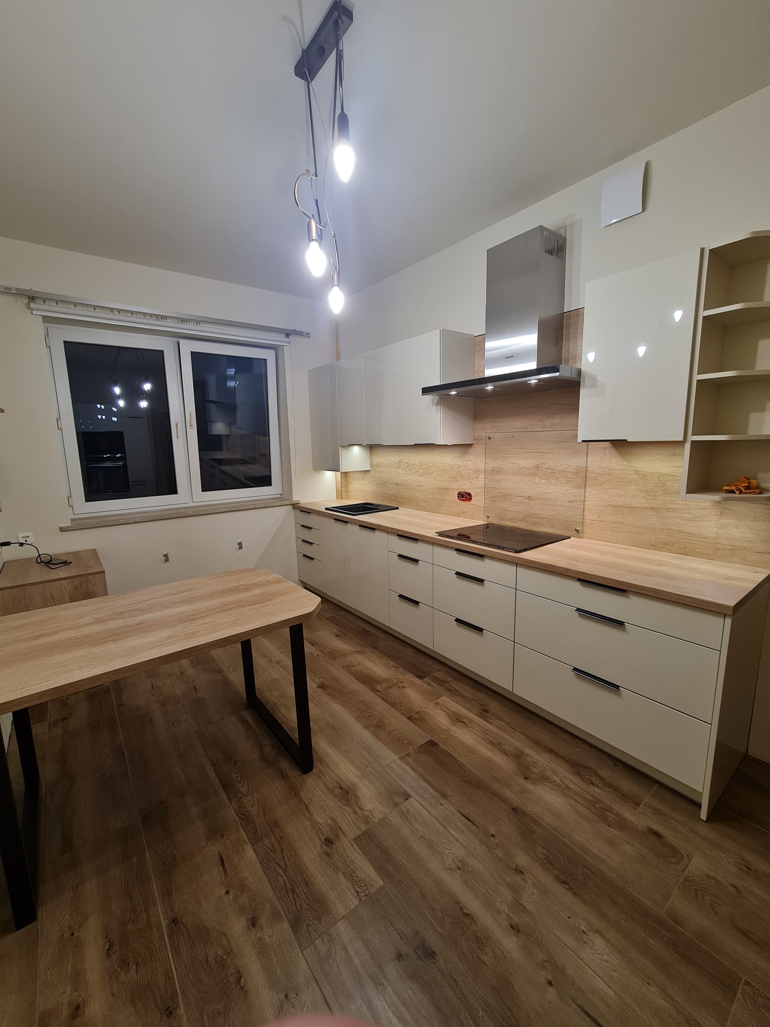 Nowoczesna kuchnia z białymi frontami szafek, drewnianym blatem i panelem ściennym o strukturze drewna. Widoczny stół z drewnianym blatem i czarnymi nogami, oraz wisząca lampa z kilkoma żarówkami.