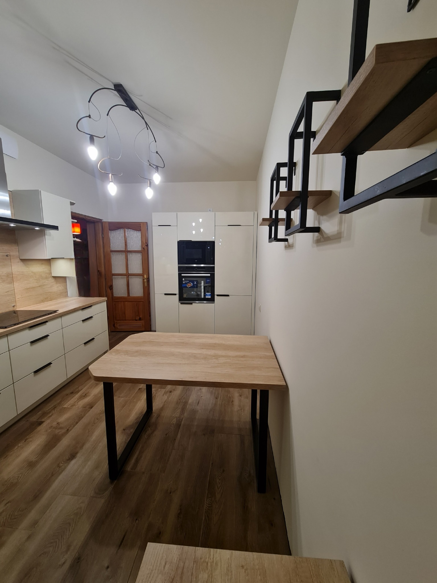 Nowoczesna kuchnia z białymi frontami, drewnianym blatem i stołem przymocowanym do ściany, oświetlona designerską lampą sufitową.