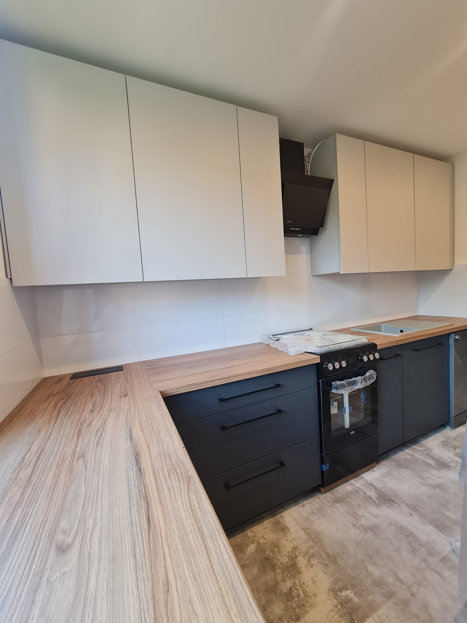 Jasna kuchnia z drewnianym blatem i czarnymi szafkami dolnymi, piekarnik w folii ochronnej, widoczny fragment podłogi.