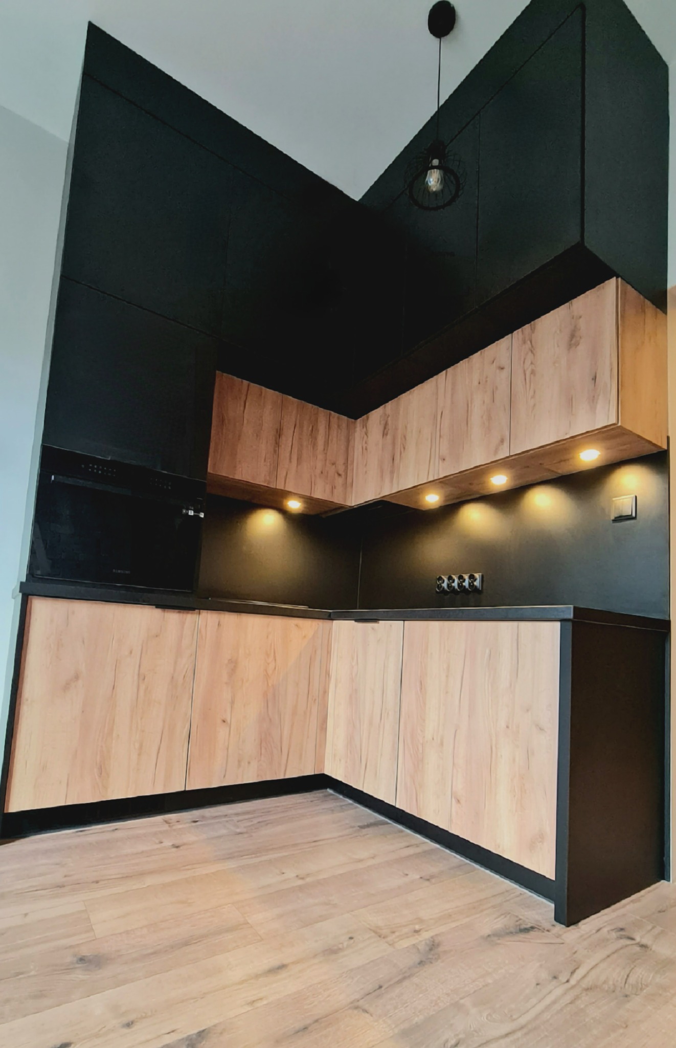 Nowoczesna kuchnia z drewnopodobnymi frontami szafek dolnych i górnych, czarne ściany i zabudowa, oświetlenie podszafkowe LED, lampa sufitowa z ozdobnym kloszem.