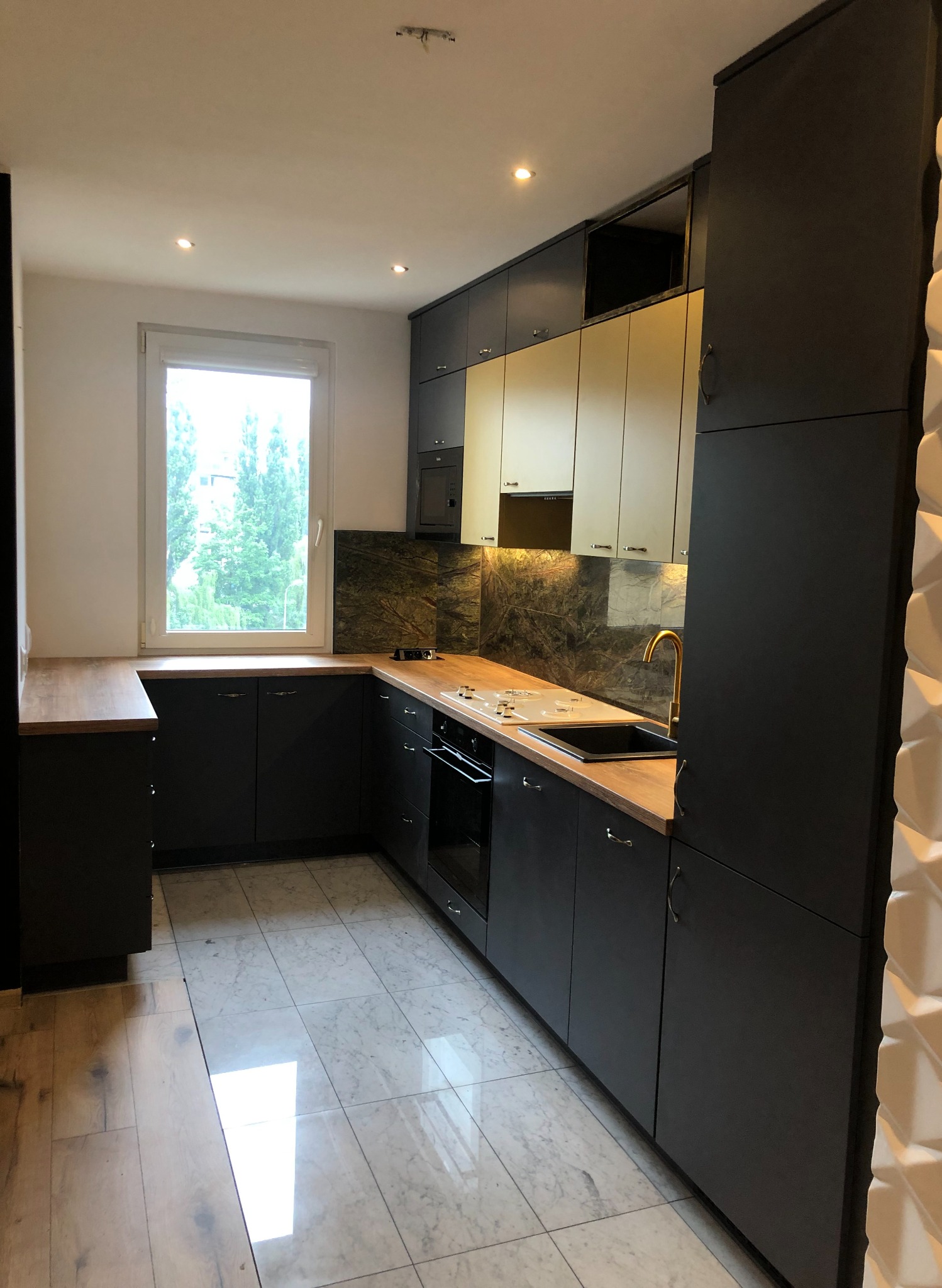 Nowoczesna kuchnia w odcieniach szarości i bieli, z drewnianym blatem i marmurowym backsplashiem, złota bateria, oświetlenie punktowe i dekoracyjna ściana z trójwymiarową fakturą.