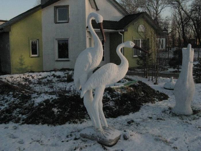 Dwie białe, betonowe figury żurawi w ogrodzie przed domem z elewacją w dwóch kolorach, z elementami śniegu.