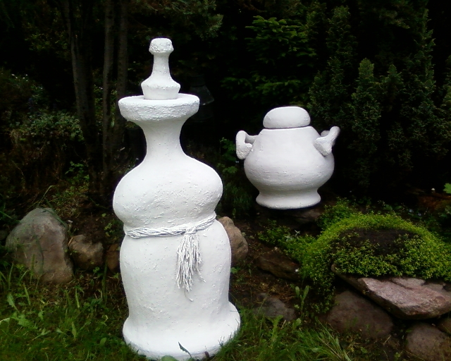 Dwie białe, ręcznie wykonane, betonowe rzeźby ogrodowe na tle zieleni: wysoka waza z dekoracyjnym sznurem i niska donica z pokrywką, ustawione na kamieniach porośniętych mchem.