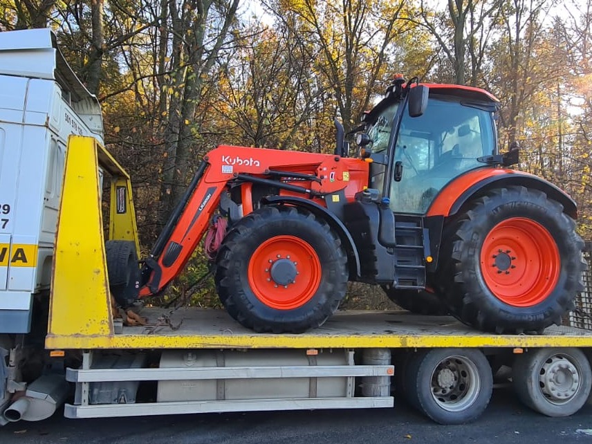 Pomarańczowy traktor Kubota z ładowaczem czołowym, transportowany na żółtej platformie lawety, widok z boku na tle drzew.