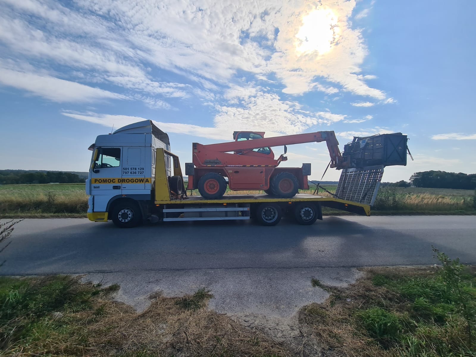 Pomoc Drogowa transportuje pomarańczowy podnośnik teleskopowy na niskopodwoziowej lawecie w słoneczny dzień.