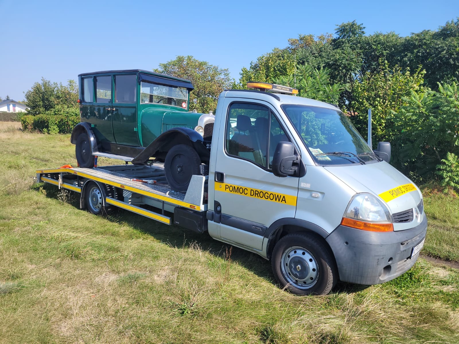 Laweta z napisem 'Pomoc Drogowa' przewozi zabytkowy, zielony samochód osobowy z czarnymi błotnikami na tle zieleni i błękitnego nieba.