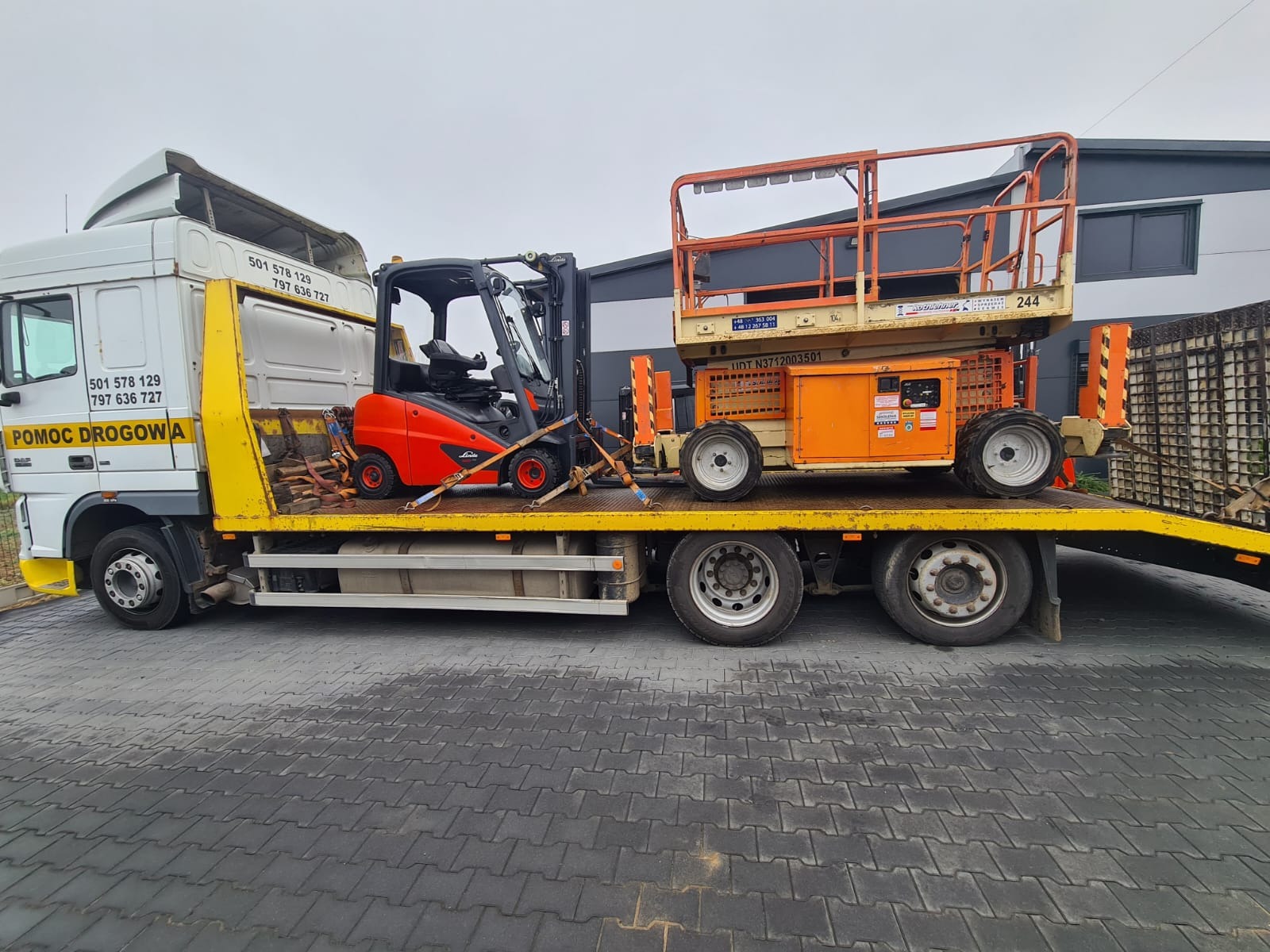 Pomoc drogowa przewozi na żółtej platformie pomarańczowy podnośnik koszowy i czerwony wózek widłowy, zabezpieczone pasami.